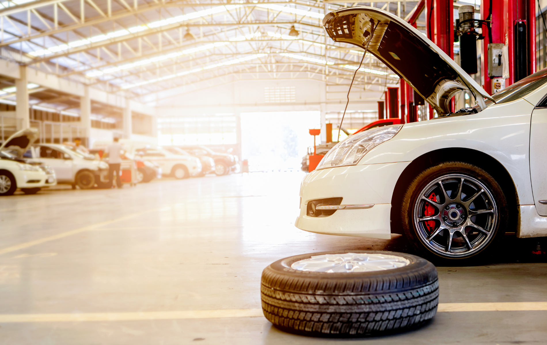 White car in auto repair shop with hood up, spare tire in foreground, other cars visible.