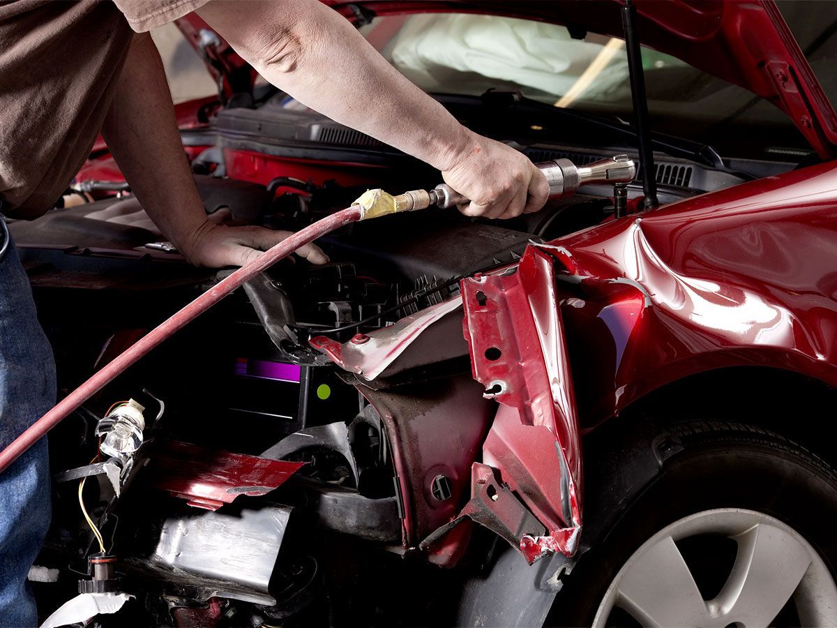 Mechanic working on a damaged red car in a repair shop, using a tool on the front end.