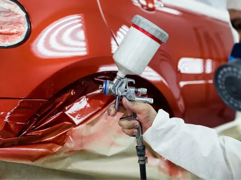Person in protective suit spraying paint onto a red car with a spray gun.