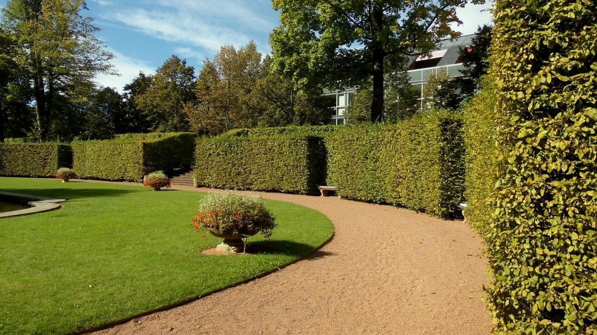 A curved gravel path winds through a formal garden with manicured hedges and green lawn under a blue sky.
