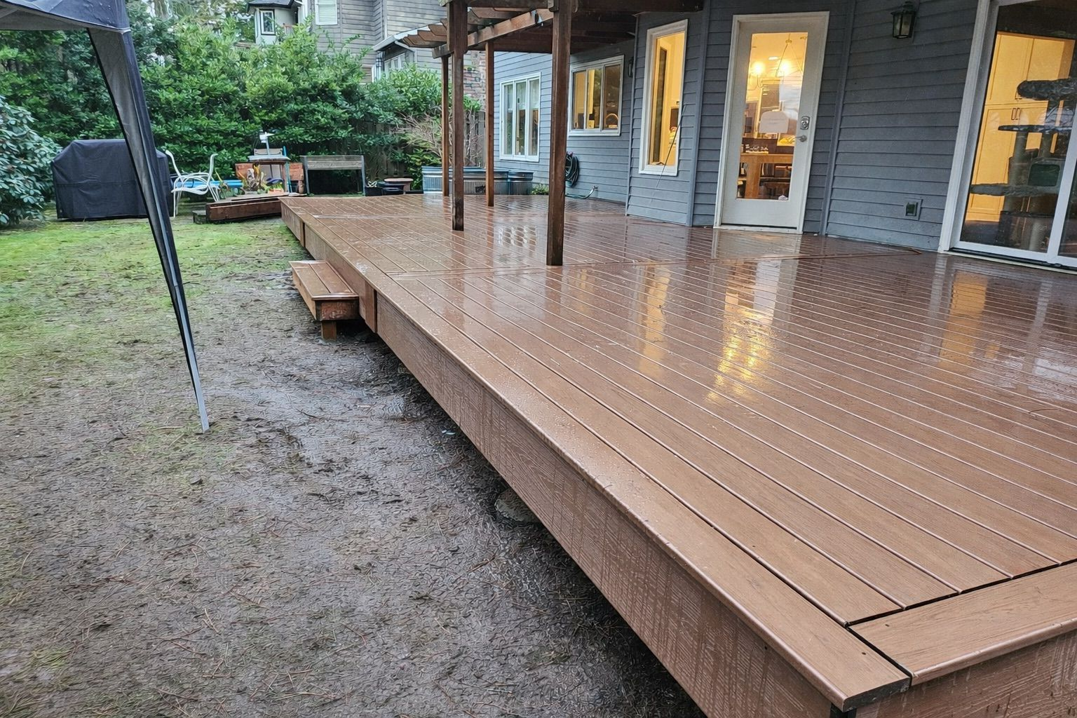 A wet, brown composite deck attached to a gray house, viewed from the side on a rainy day over a muddy lawn.