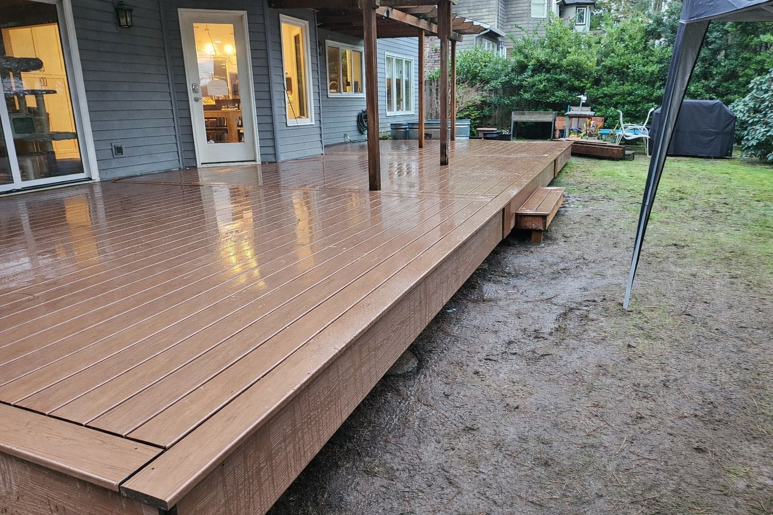 A wide-angle view of a newly installed, wet brown composite deck attached to the back of a grey-sided house.