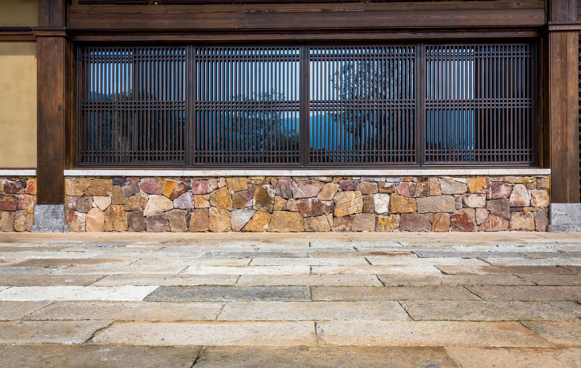 A view of a building with a large wooden-latticed window above a rustic stone foundation and a paved stone courtyard.