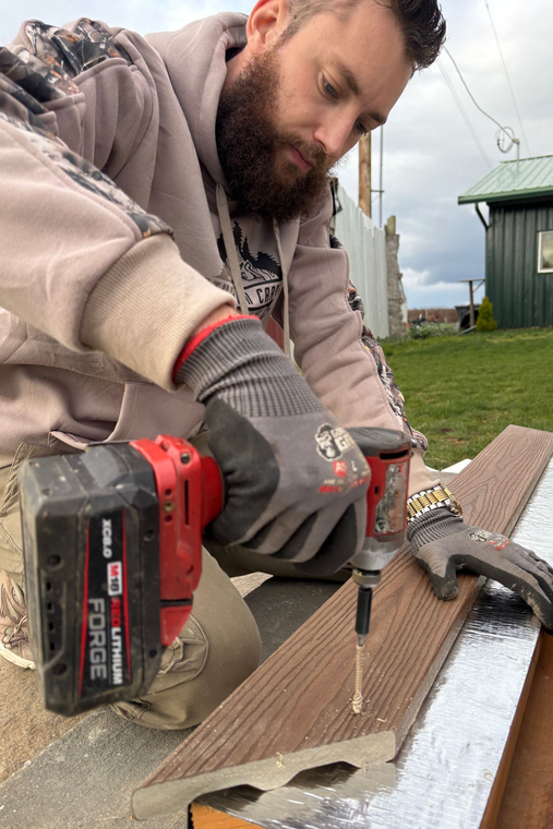 A person wearing gray work gloves uses a red cordless drill to drive a screw into a wooden board outdoors.
