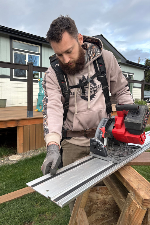 A person wearing a hoodie and a harness operates a red track saw on a guide rail, set up on a wooden sawhorse outdoors.