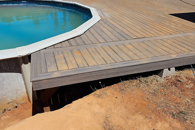 A wooden deck platform surrounding a section of an above-ground pool, elevated above bare dirt ground.