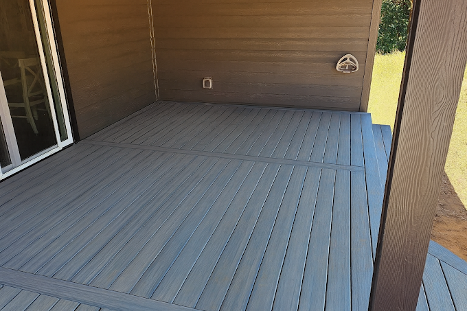 A wooden deck with grey, horizontal planks extending from a sliding glass door against a brown-sided house.