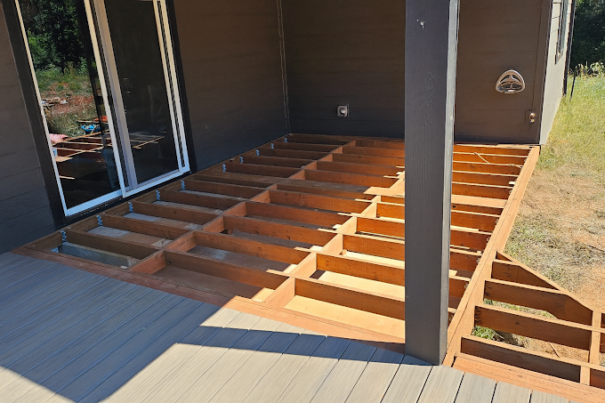 Wooden deck frame under construction outside a sliding glass door, with finished grey composite boards in the foreground.