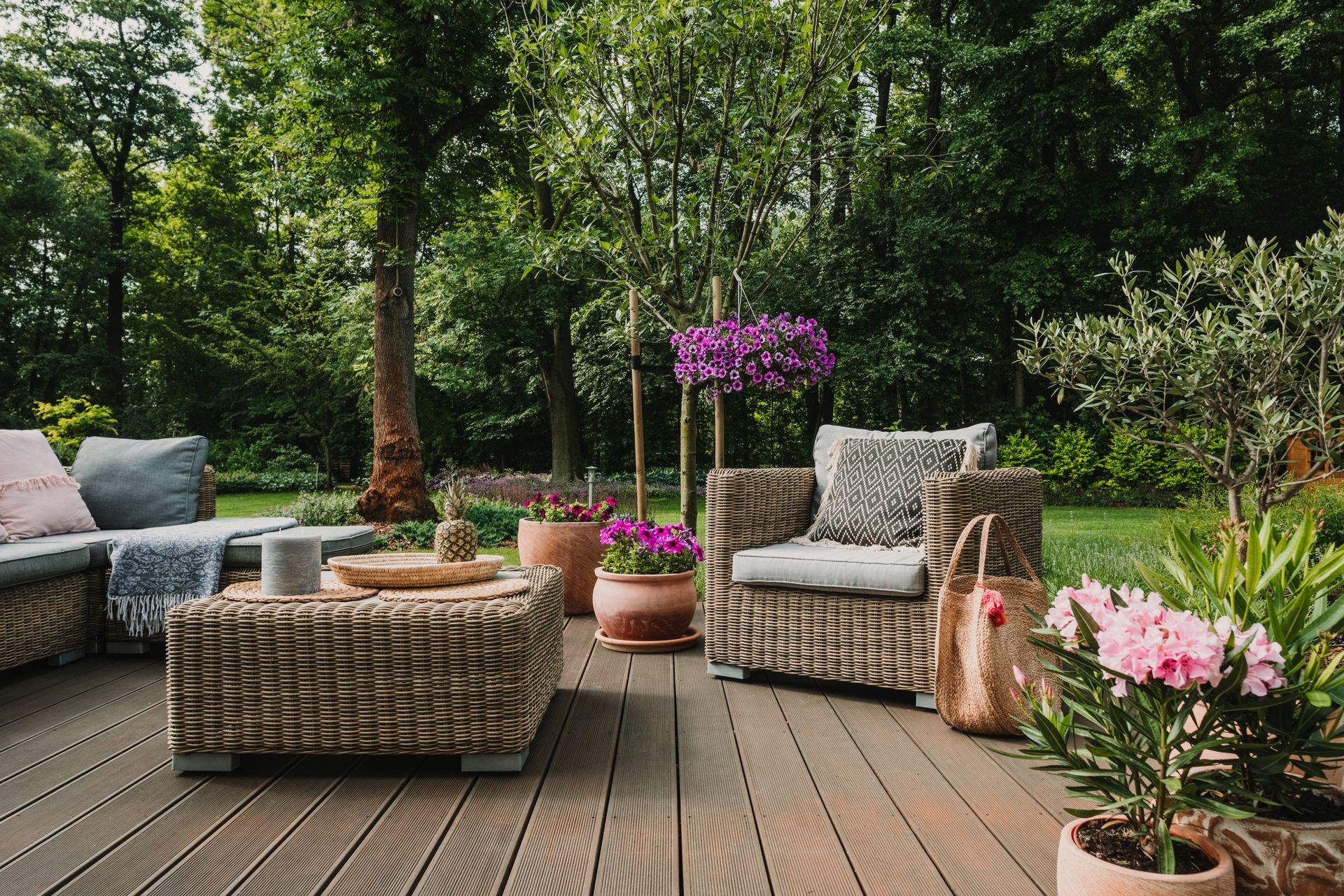 Outdoor patio seating with wicker chairs, a coffee table, and potted pink flowers beside a wooded garden.