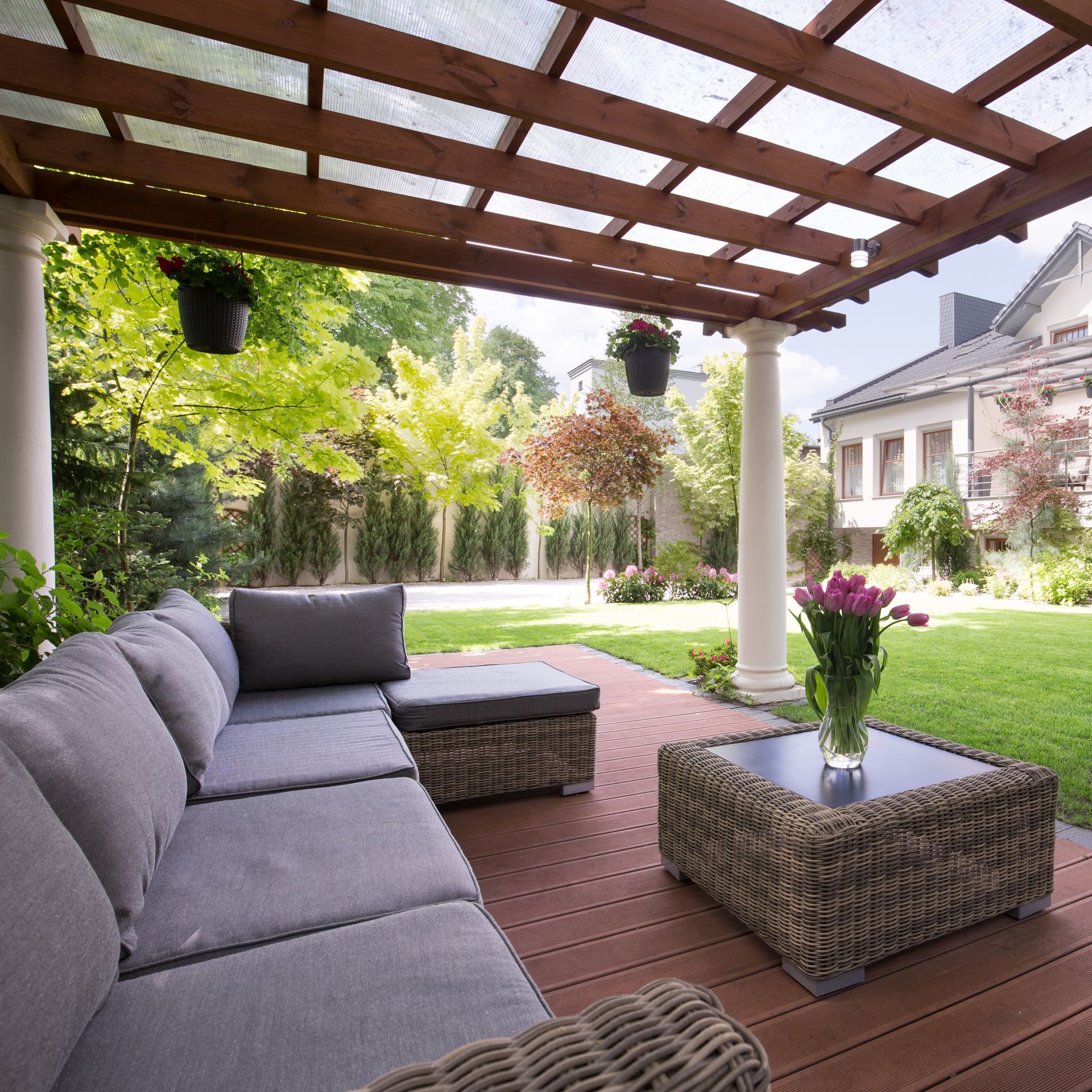 A gray sectional sofa and matching coffee table with tulips on a wooden patio under a wooden pergola, overlooking a yard.
