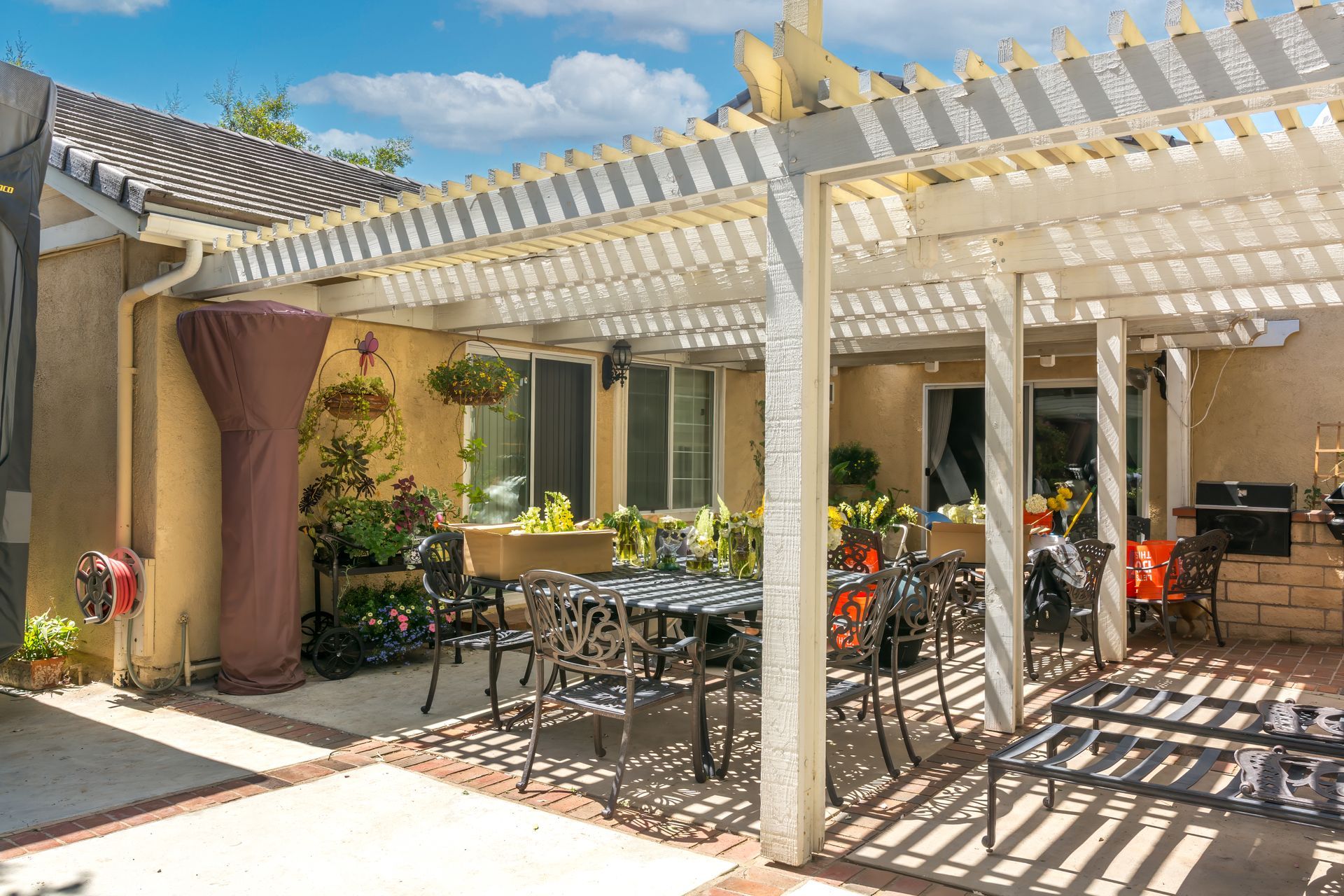 A white wooden pergola covers a patio area with a dining set, potted plants, and outdoor furniture in a backyard setting.