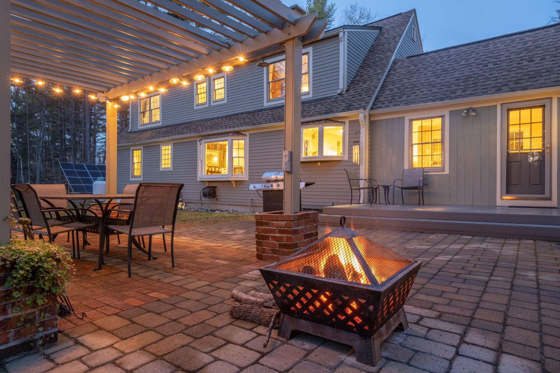 A lit fire pit sits on a stone patio in front of a gray house with a pergola and outdoor dining area at dusk.
