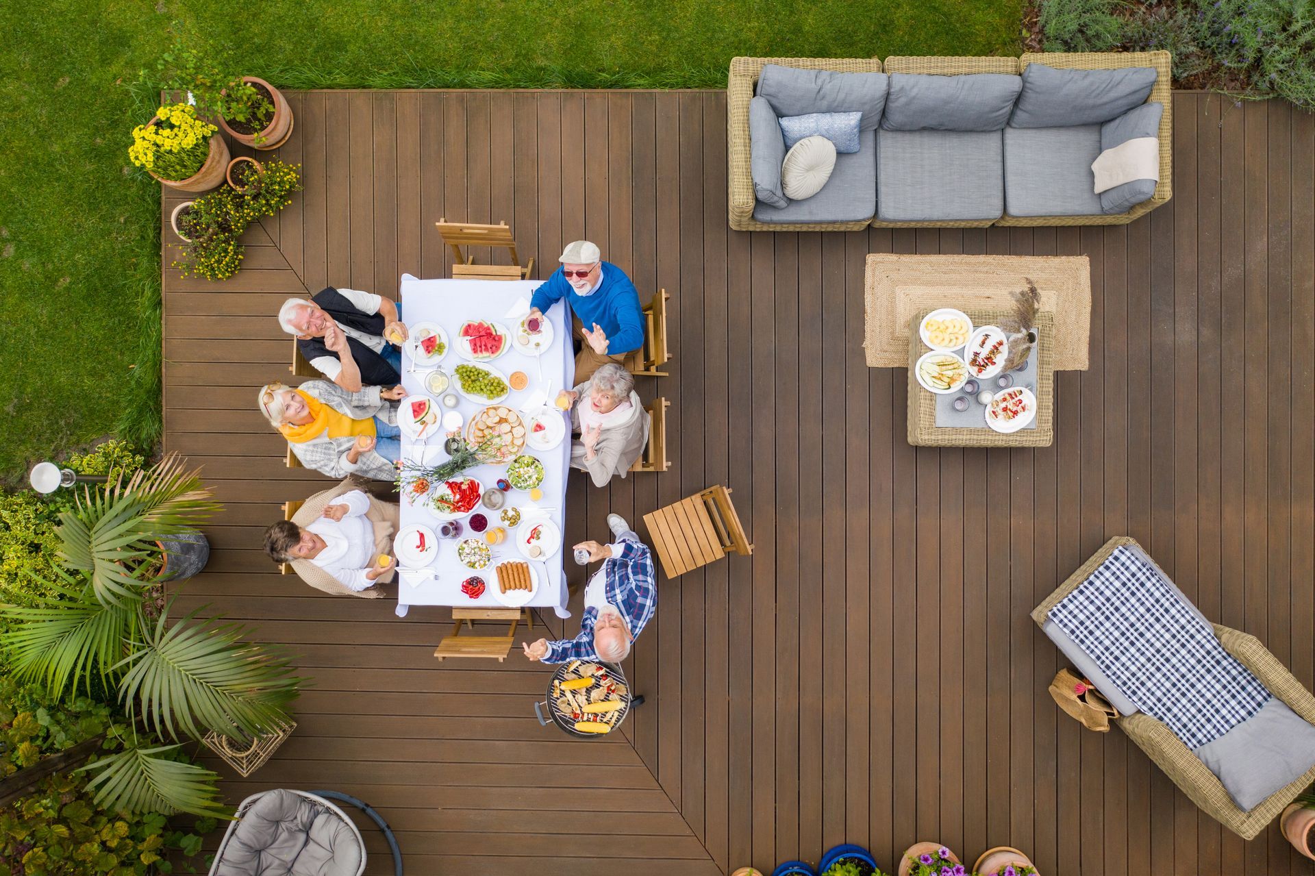 An overhead view of people eating at a rectangular table on a wooden deck, with outdoor seating and plants nearby.