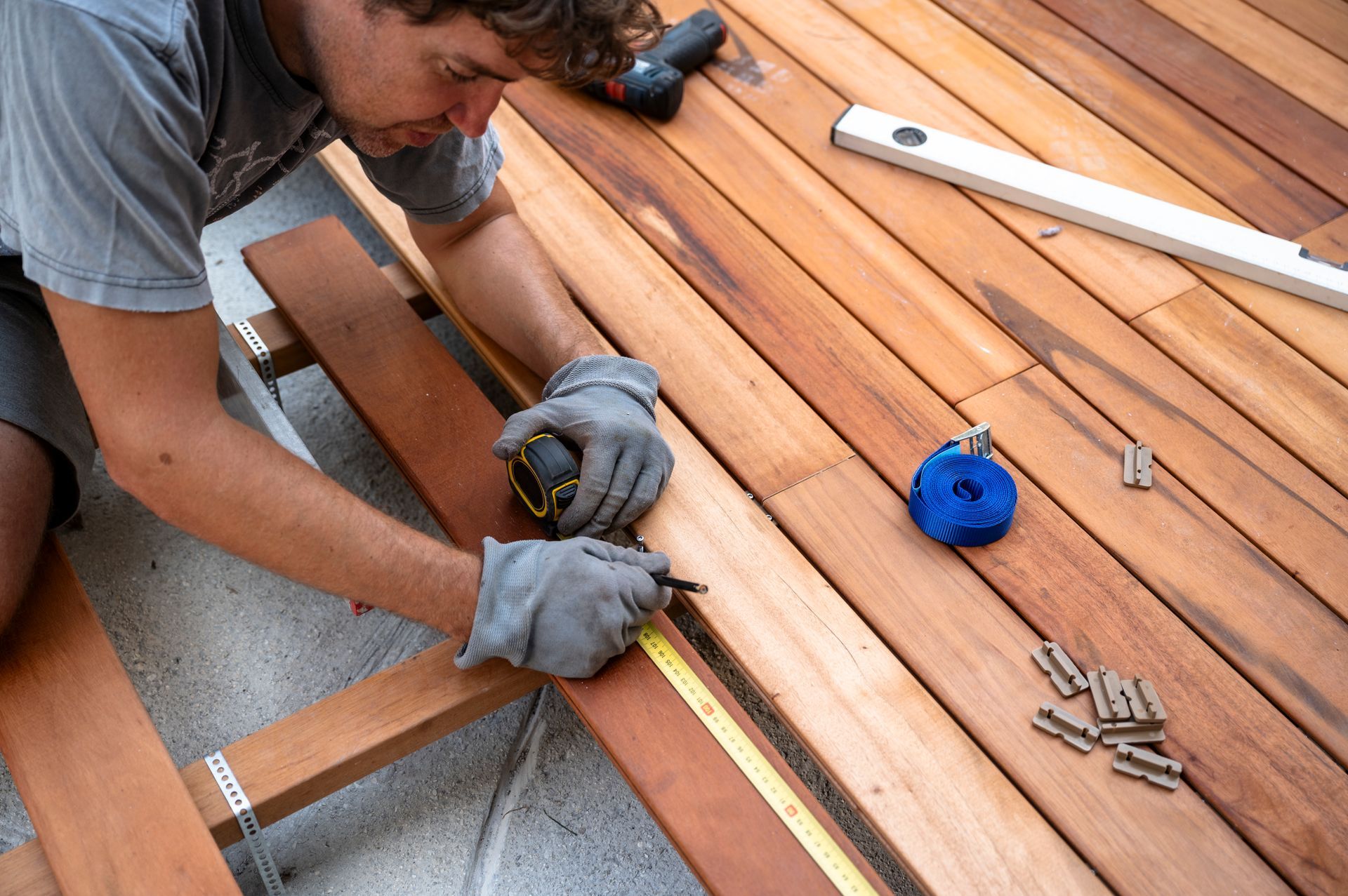 A person wearing grey work gloves measures and marks a wooden deck plank with a tape measure and pencil.