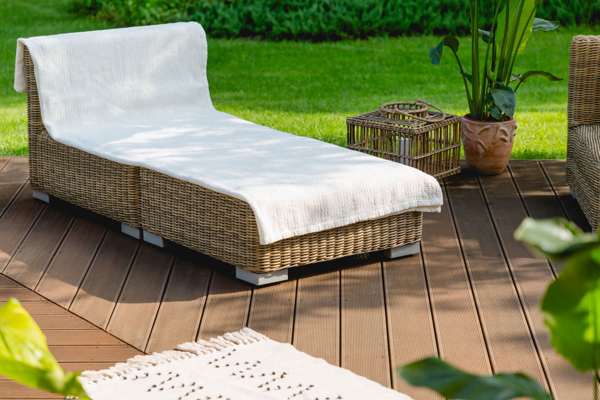 A rattan lounge chair with a white blanket sits on a wooden deck in a garden.