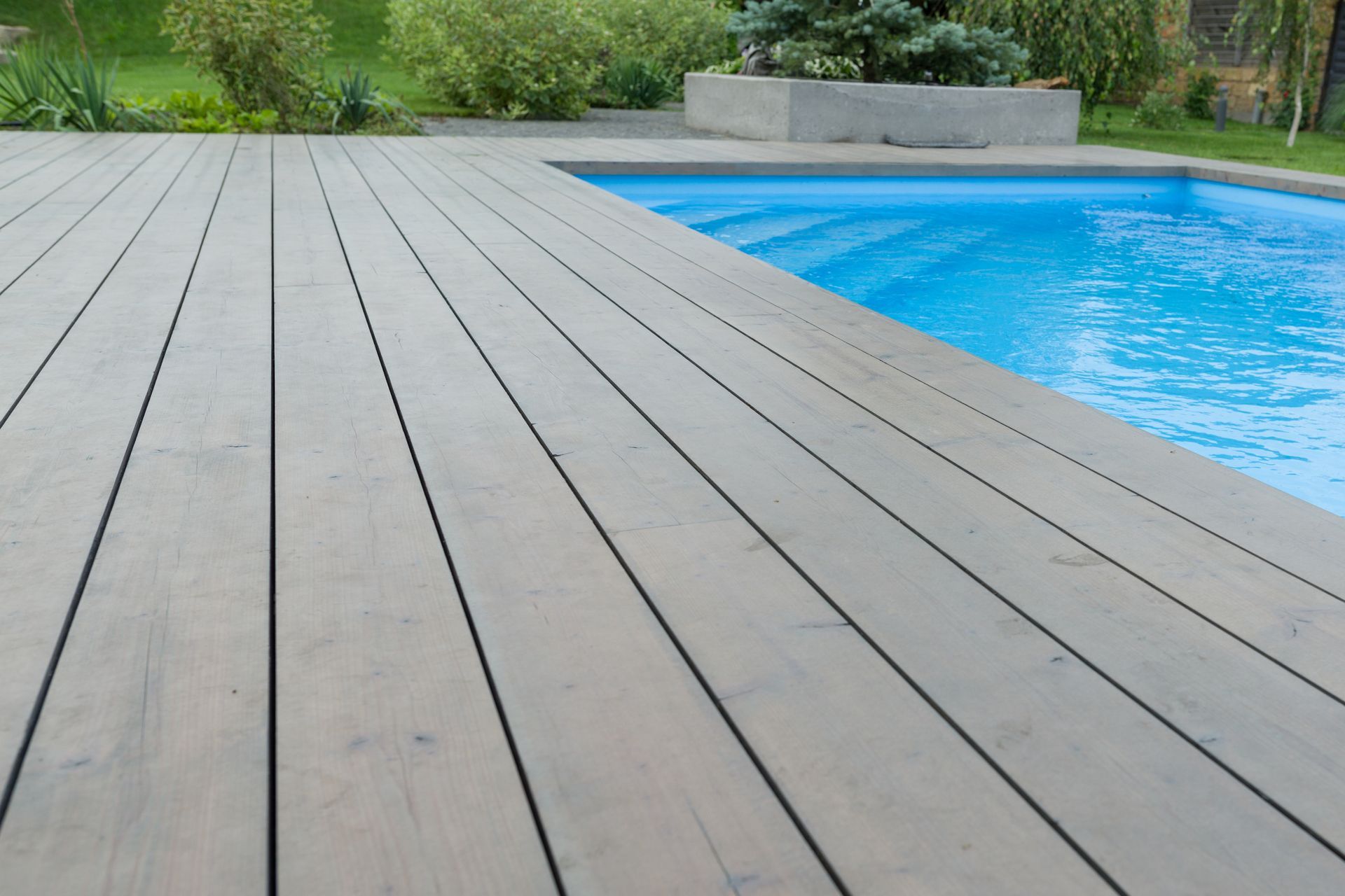 Modern gray deck boards beside a bright blue swimming pool, with green landscaping in the background