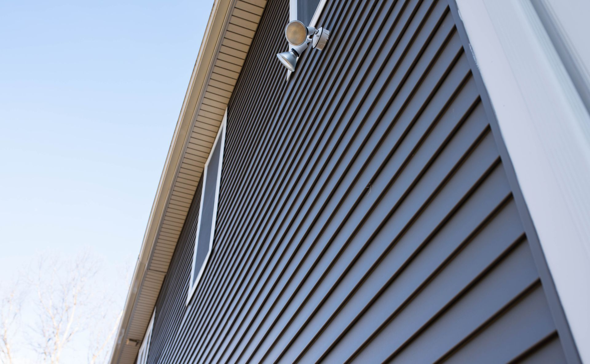 A low-angle view of a grey-sided house with a white trim eave, a visible window, and a mounted outdoor security light.