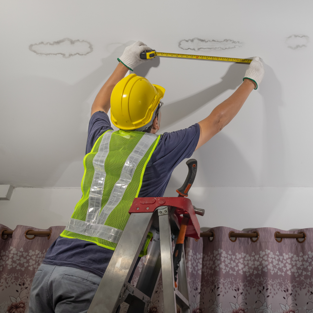 A man is measuring a water damage spot on a wall with a tape measure.