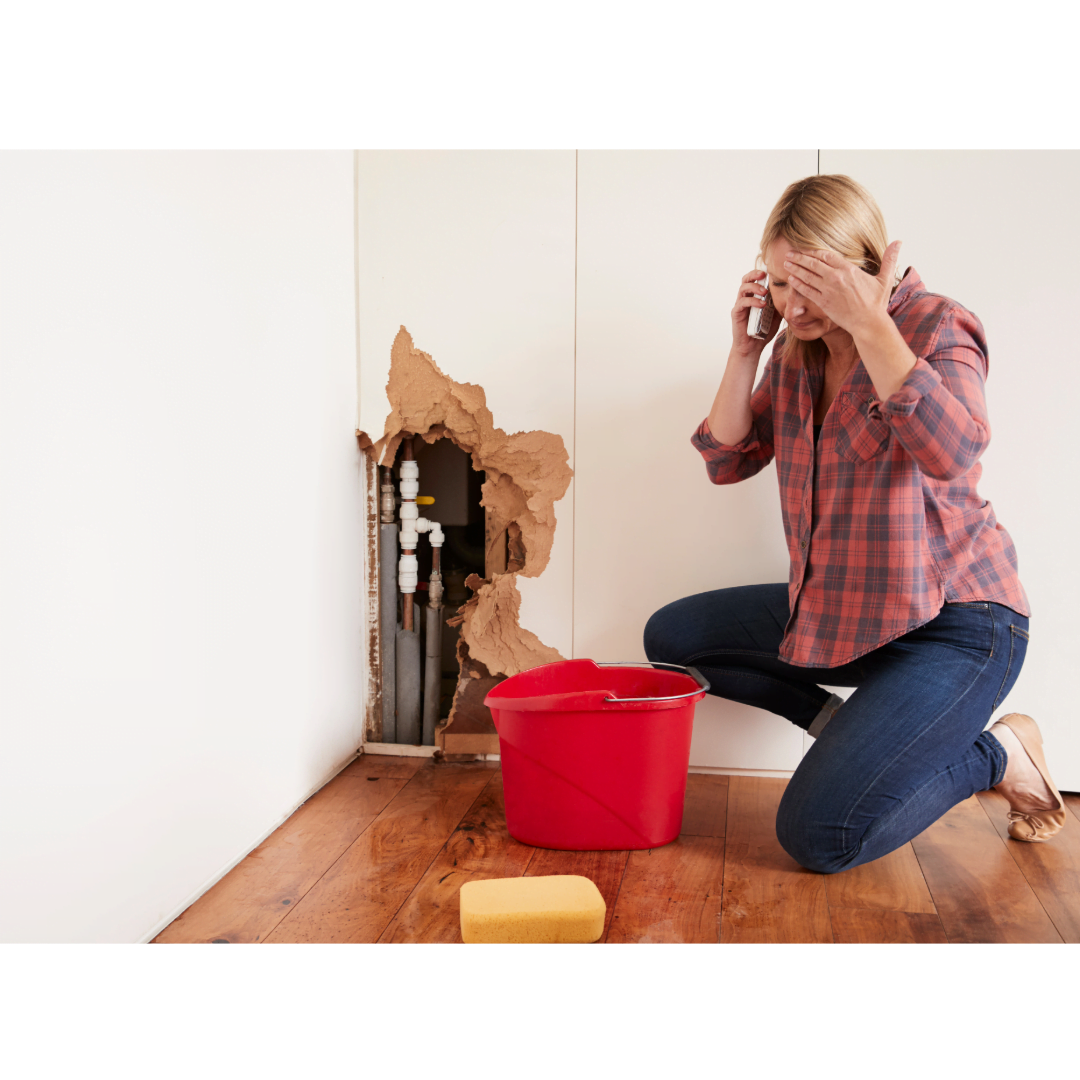 A woman is kneeling down next to a red bucket and a hole in the wall and talking on a cell phone.