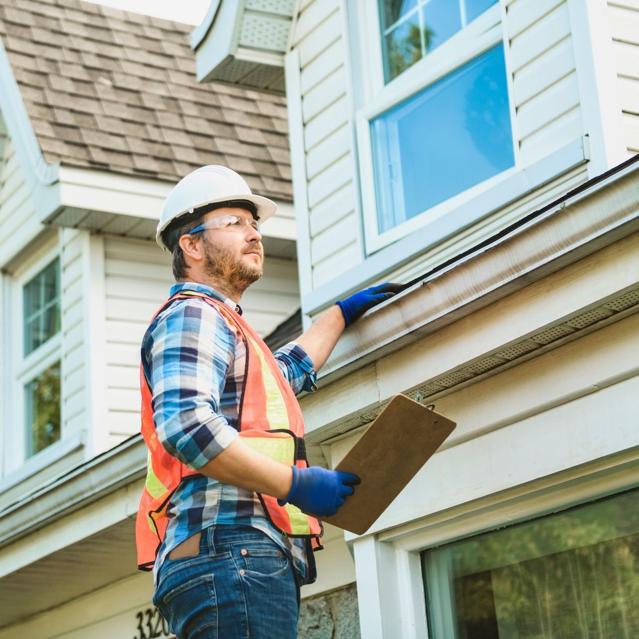 A man wearing a hard hat and safety vest is looking out a window while holding a clipboard.