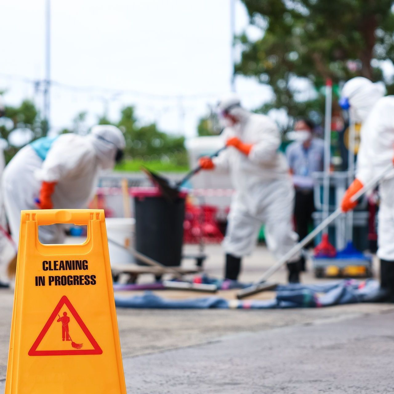 A yellow caution sign that says cleaning in progress and people in protective suits cleaning behind it