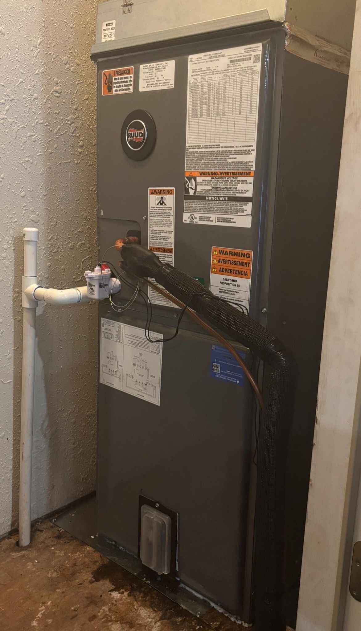 A grey HVAC unit in a room with a white wall and pipe, with warning labels and black insulated wires.