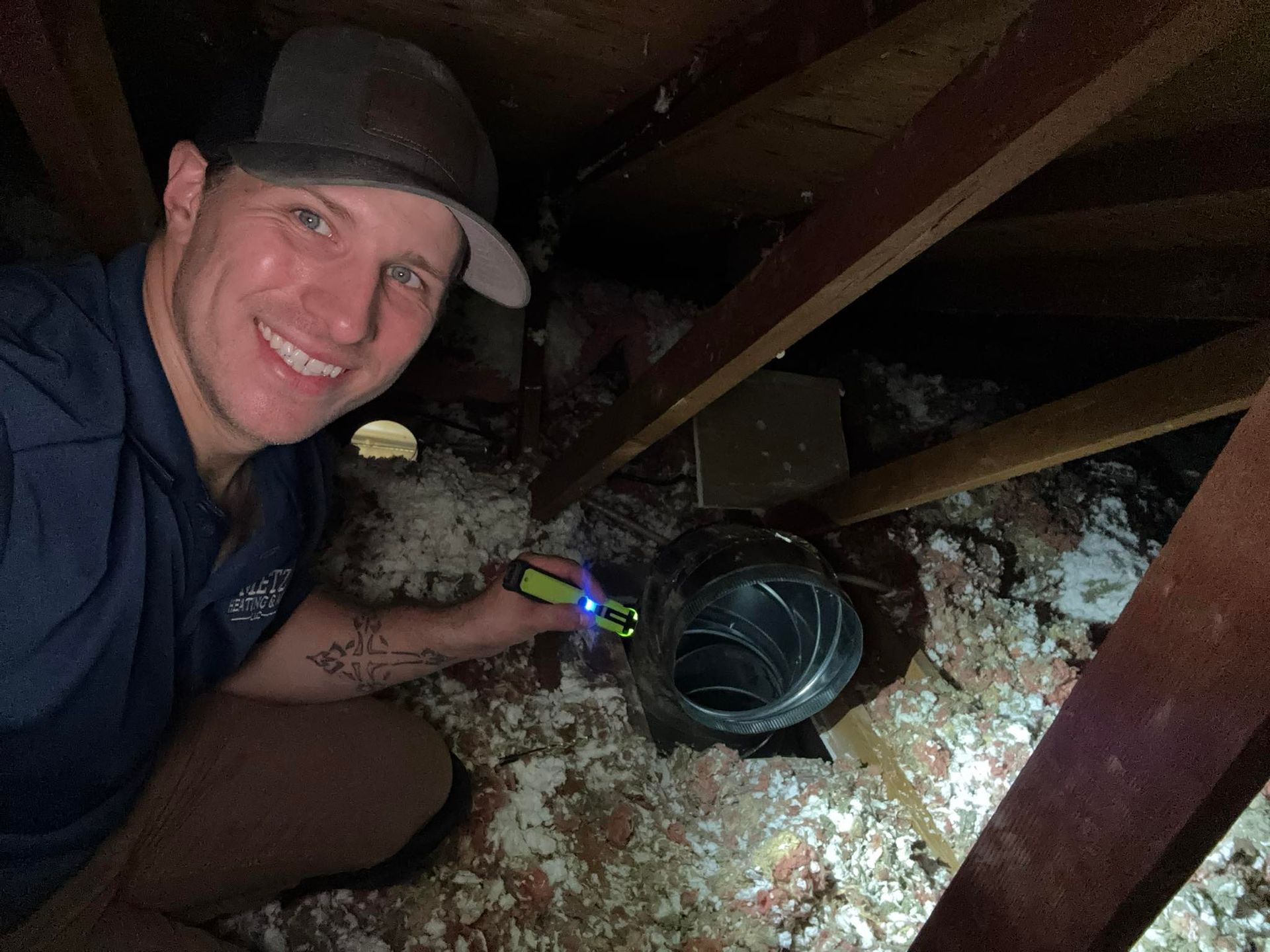 A man smiles while holding a flashlight in an attic inspecting ductwork.