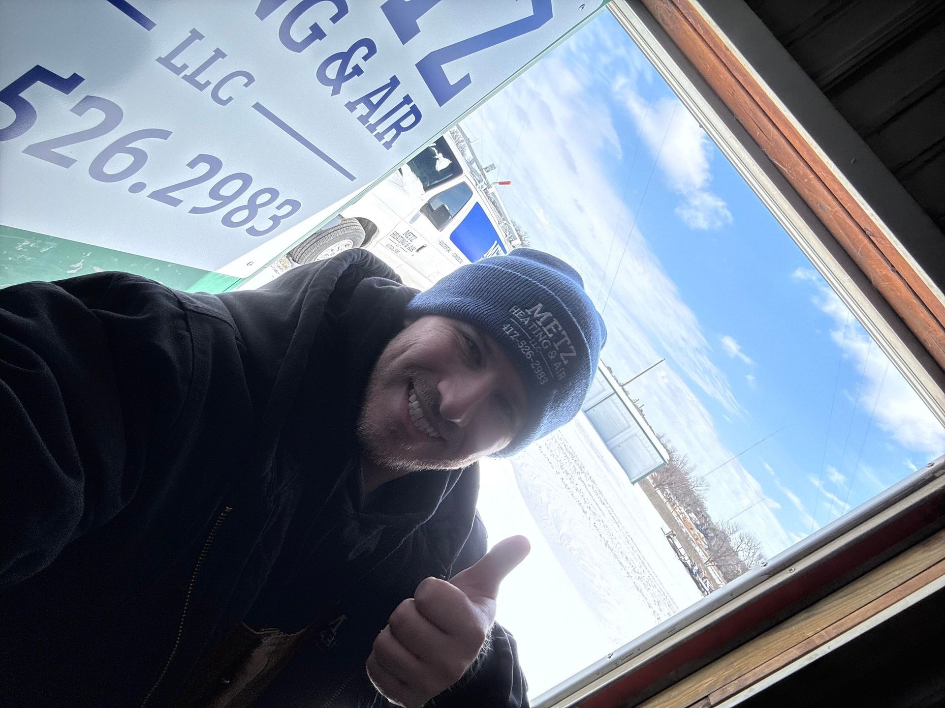Man in beanie gives thumbs up near window; company sign, snowy outdoors.