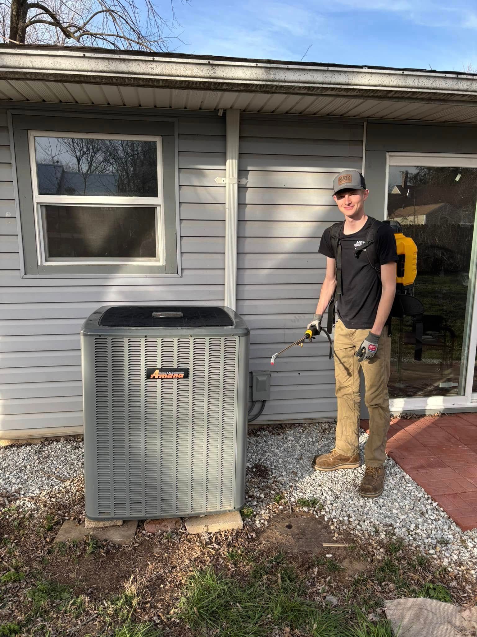 Man standing next to an air conditioning unit outside a house, holding a sprayer.