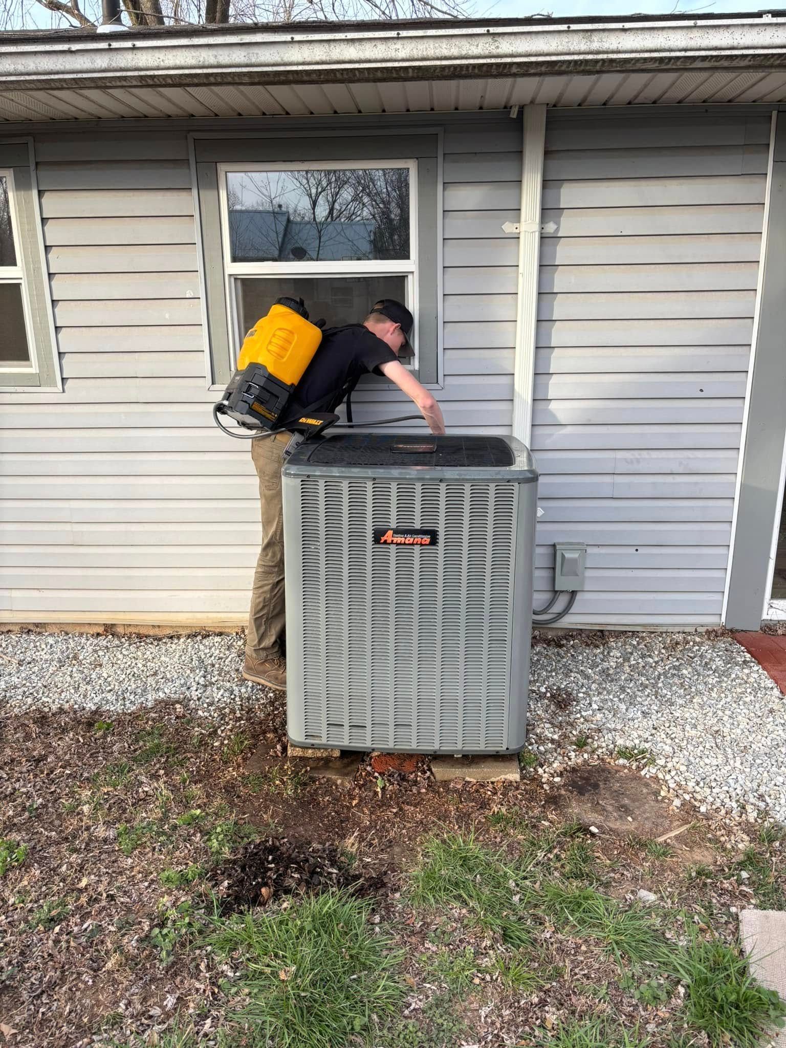 Person sprays AC unit outside a building with gray siding, next to a window.