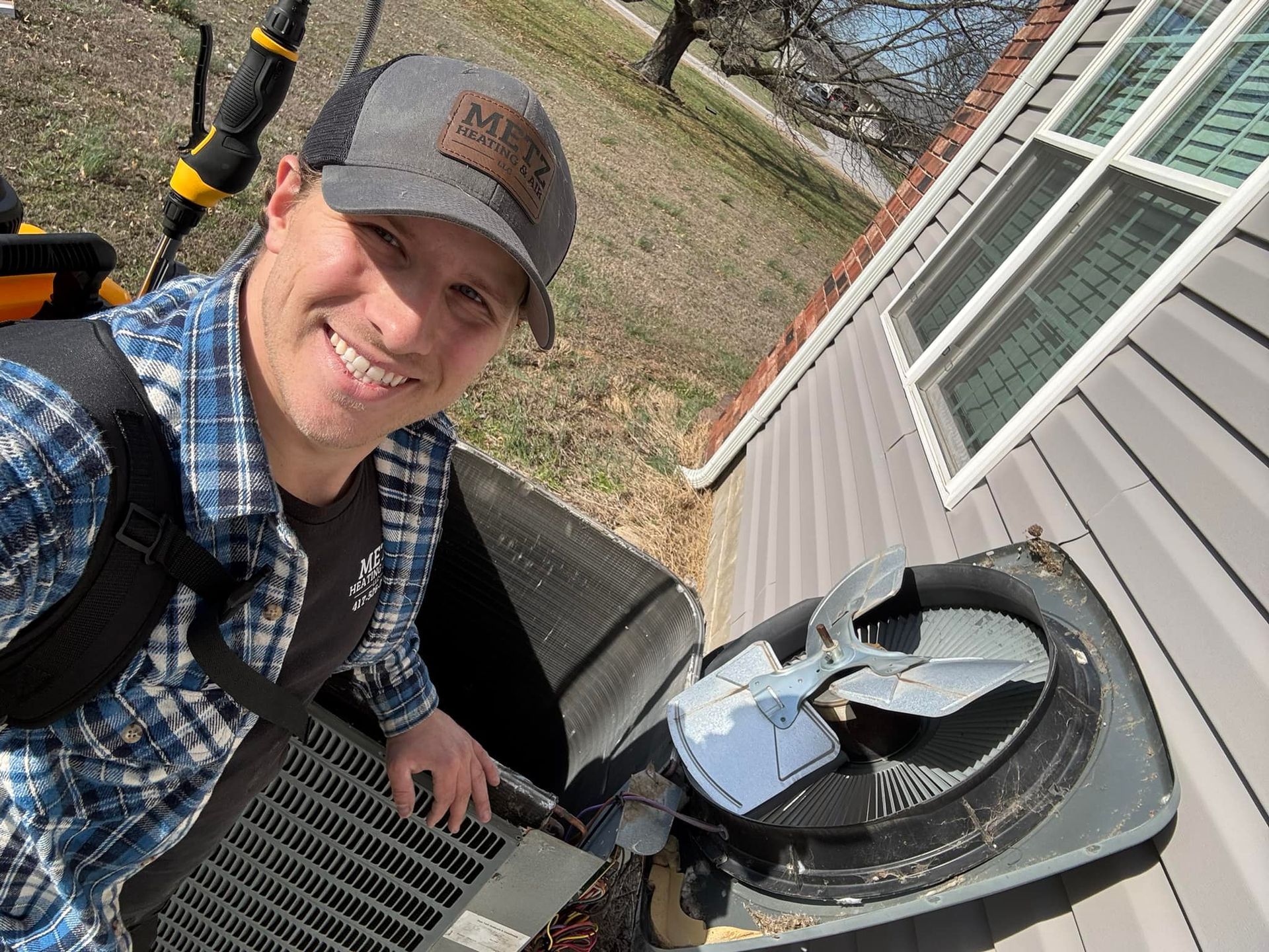 Man smiles at camera next to an open air conditioning unit. Gray and blue, outdoors.