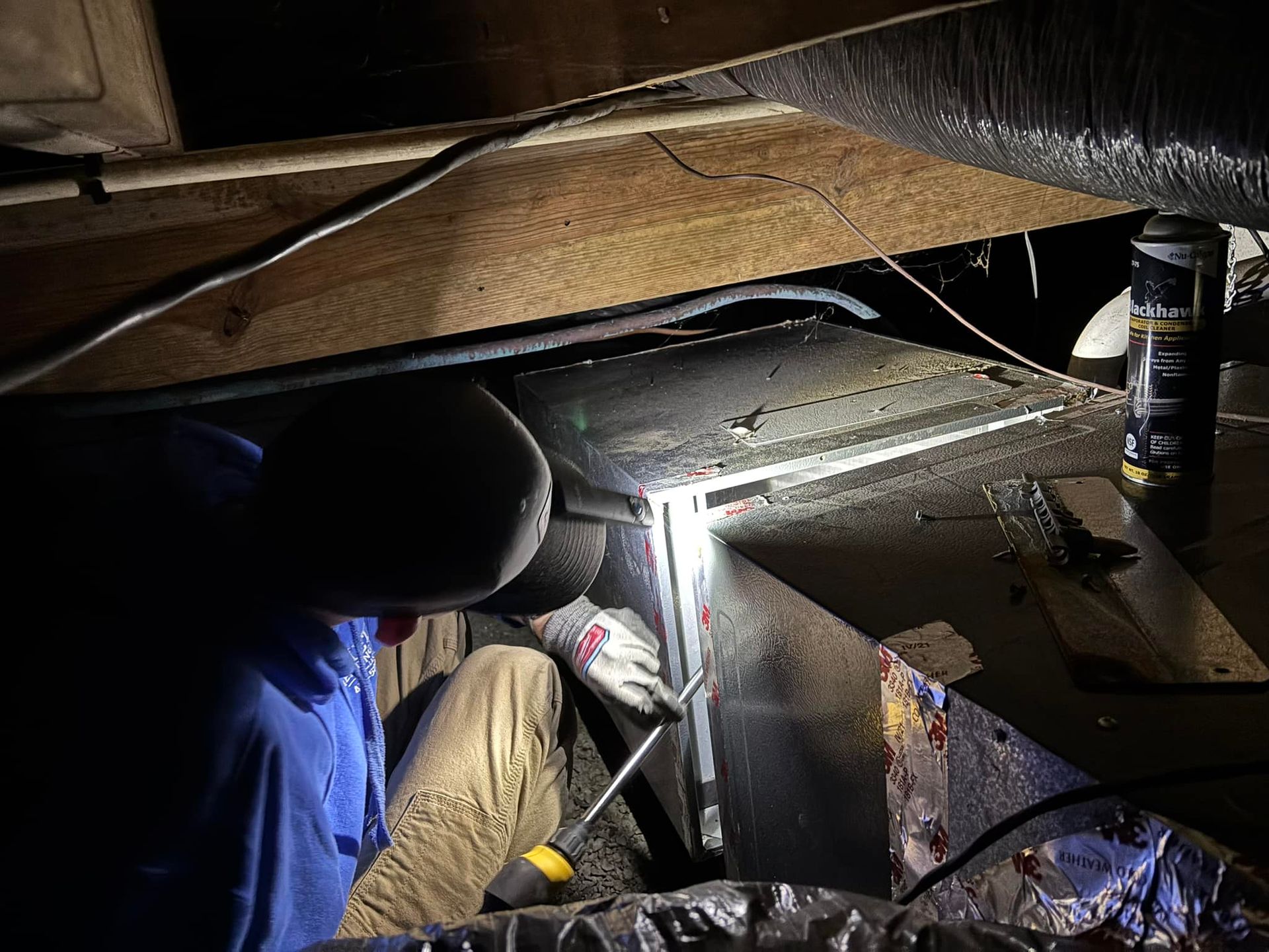 Person in blue shirt working on HVAC ductwork in a dim attic.