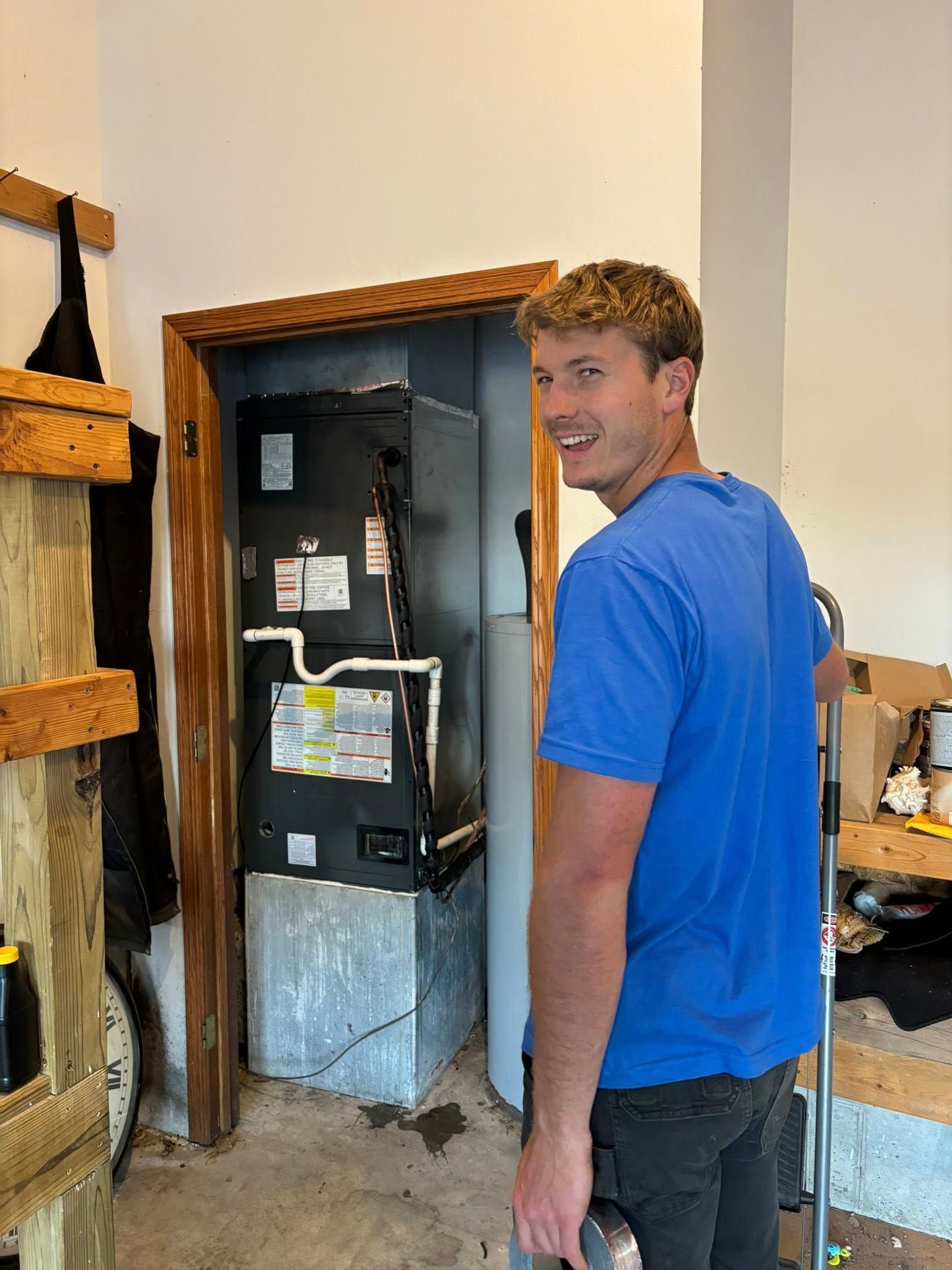 Man smiles near furnace in a doorway. Blue shirt, gray metal base, wooden frame. Garage setting.