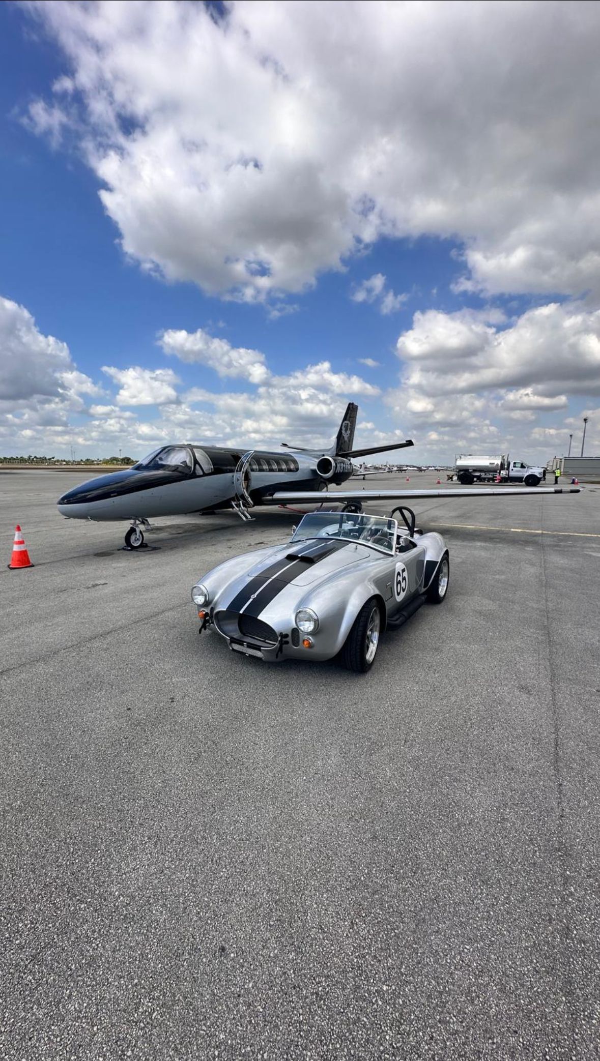 A silver sports car is parked next to an airplane on a runway.