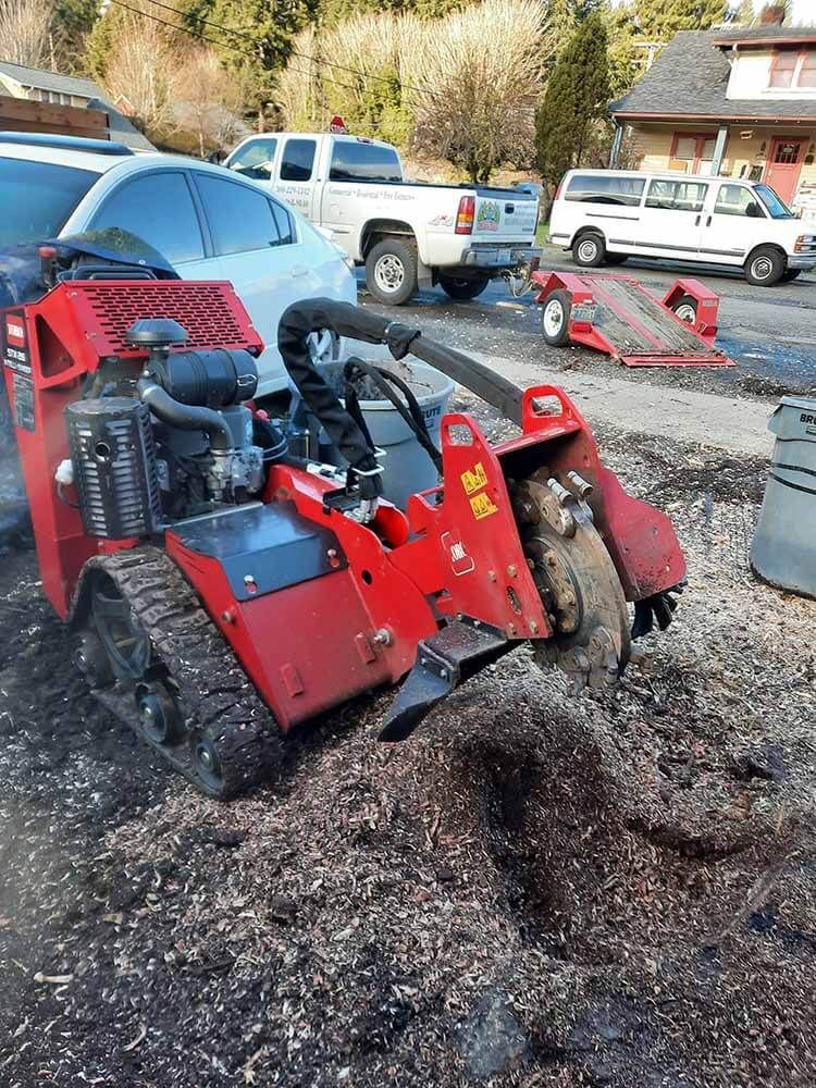 A red stump grinder is cutting a tree stump in a parking lot.