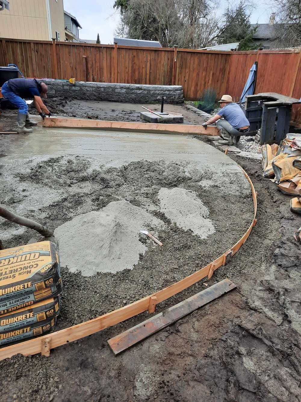 Two men are working on a concrete driveway in a backyard.