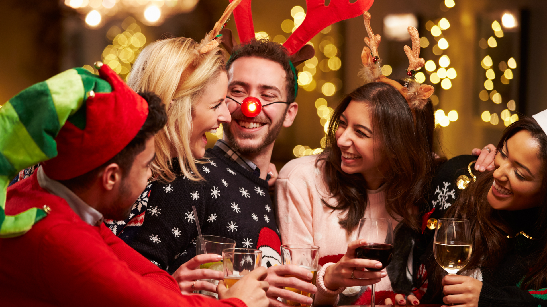 Image with five friends dressed in Christmas attire. They have alcoholic drinks in their hands and are smiling and laughing while looking at each other.