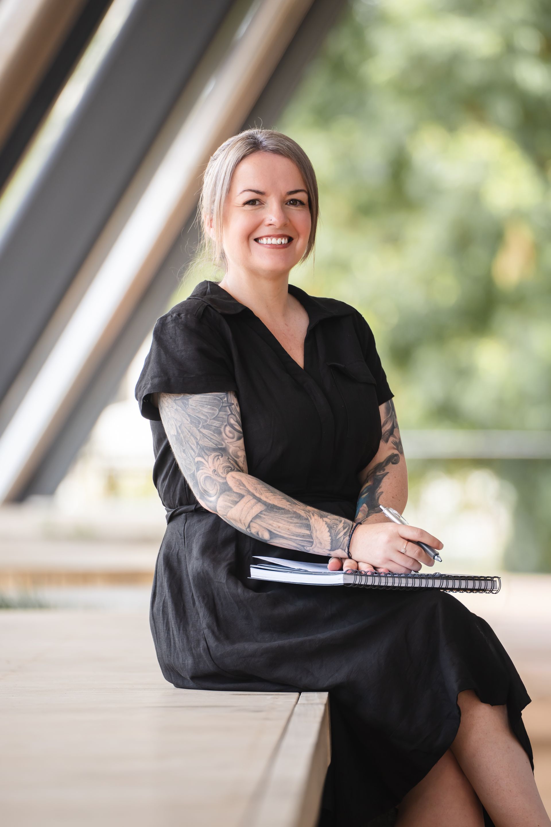 Woman with tattoos, smiling, holding notebook and pen, sitting outdoors.