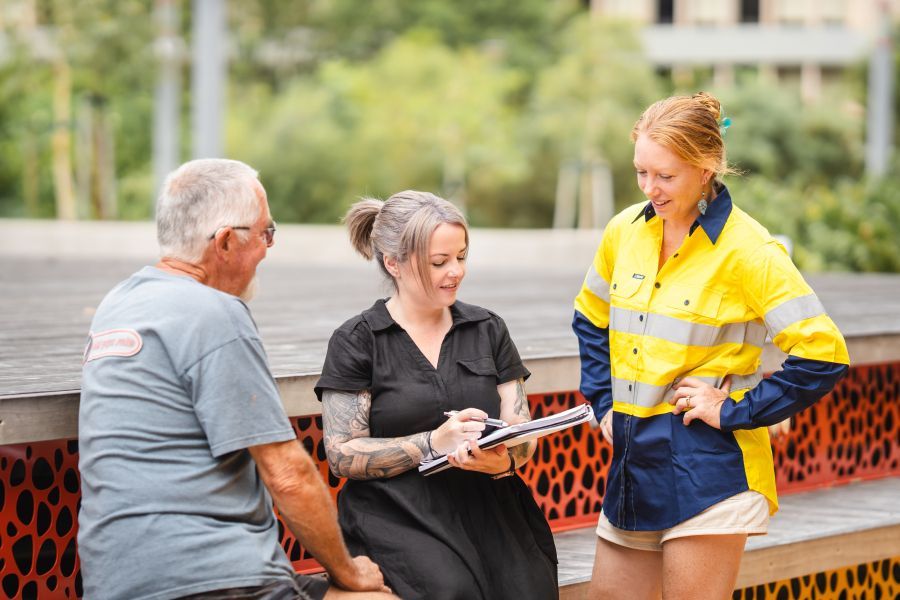 Three people sitting and talking outdoors: one man, two women, one writing on a clipboard.