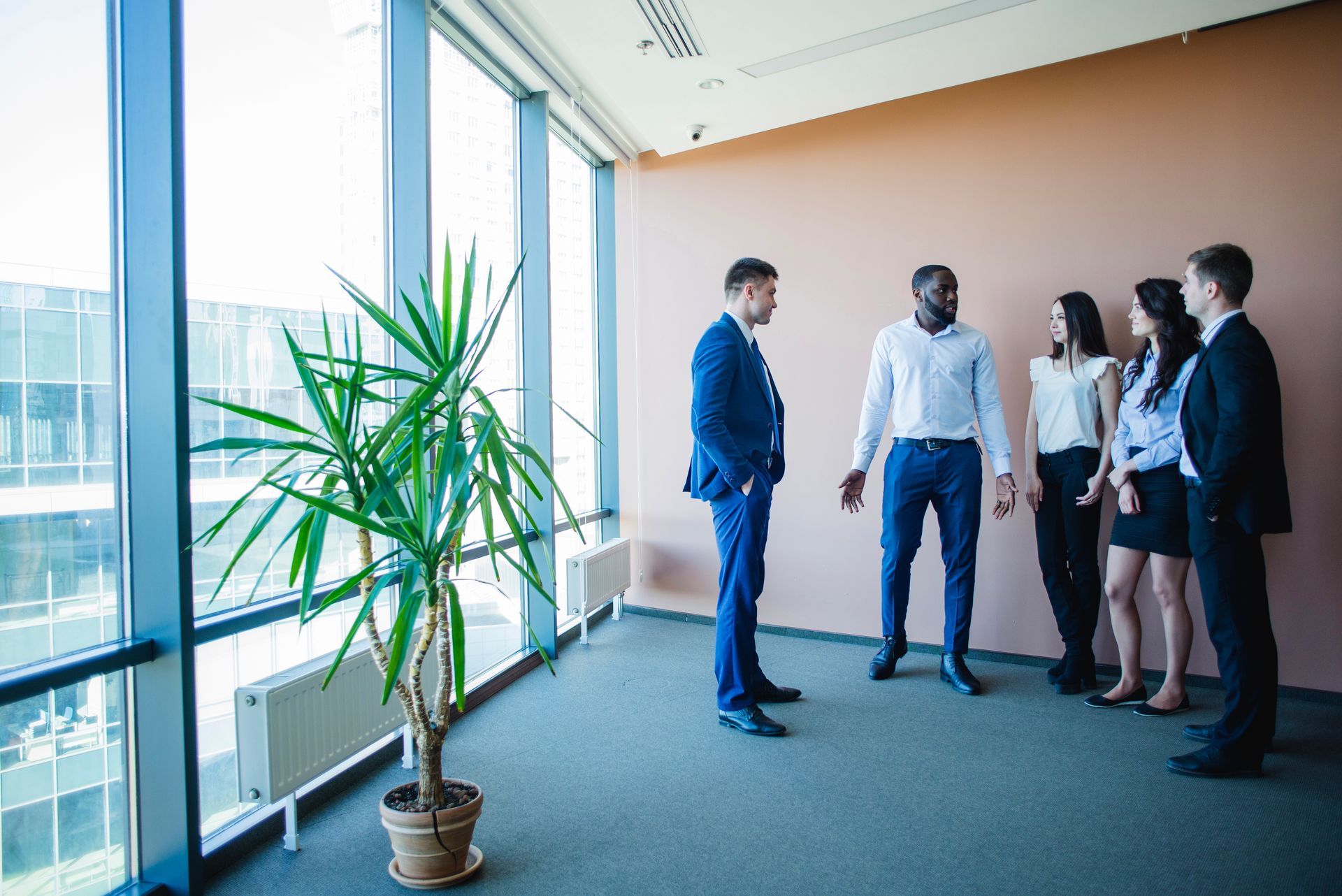 Business meeting in a modern office. Men and women in suits stand near a large window, listening to two men.