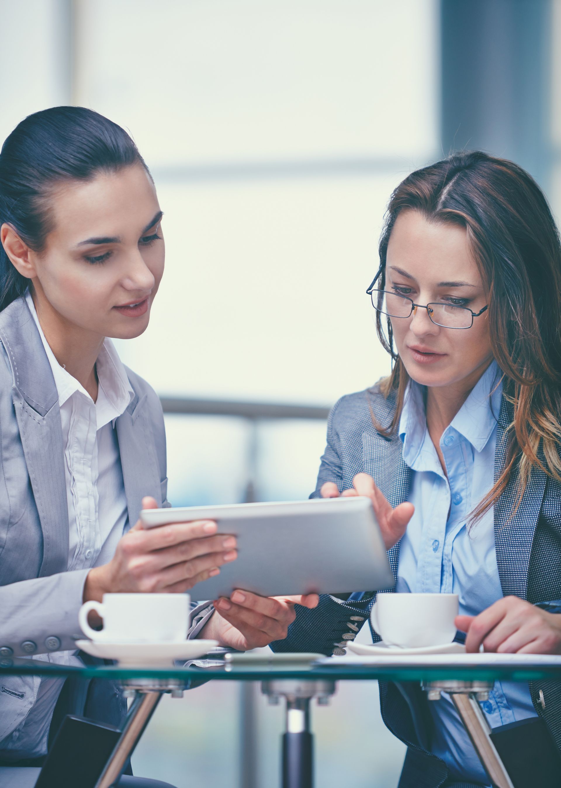 Two women looking at tablet, sitting at table with coffee cups. Office setting.