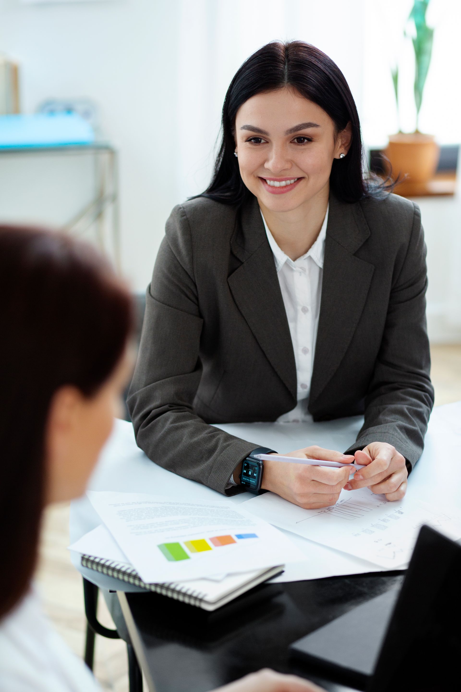 Woman in blazer smiles, meeting over documents with another person at a table in an office.