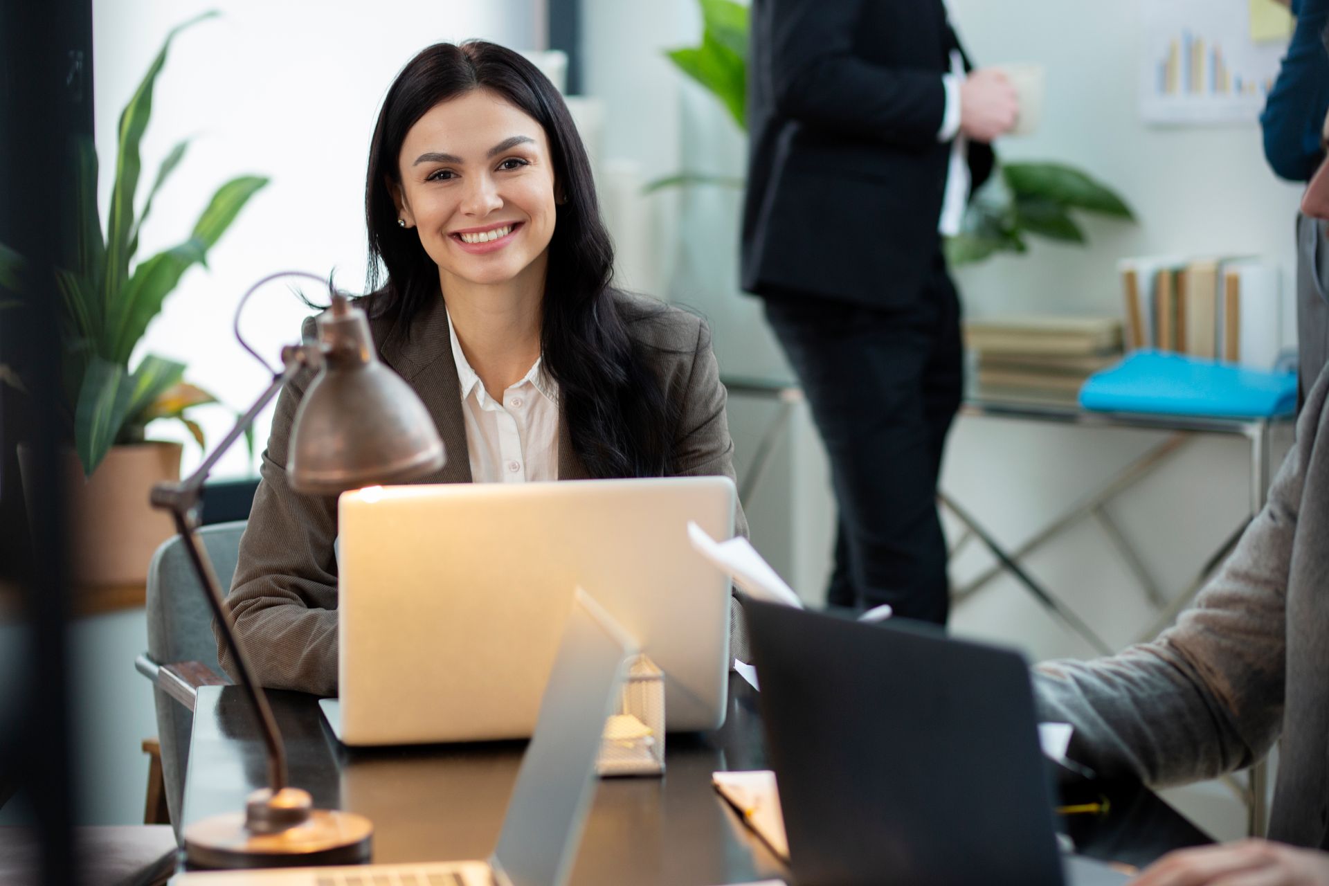 Woman smiles, working on laptop at desk; coworkers in background. Office setting with plants.