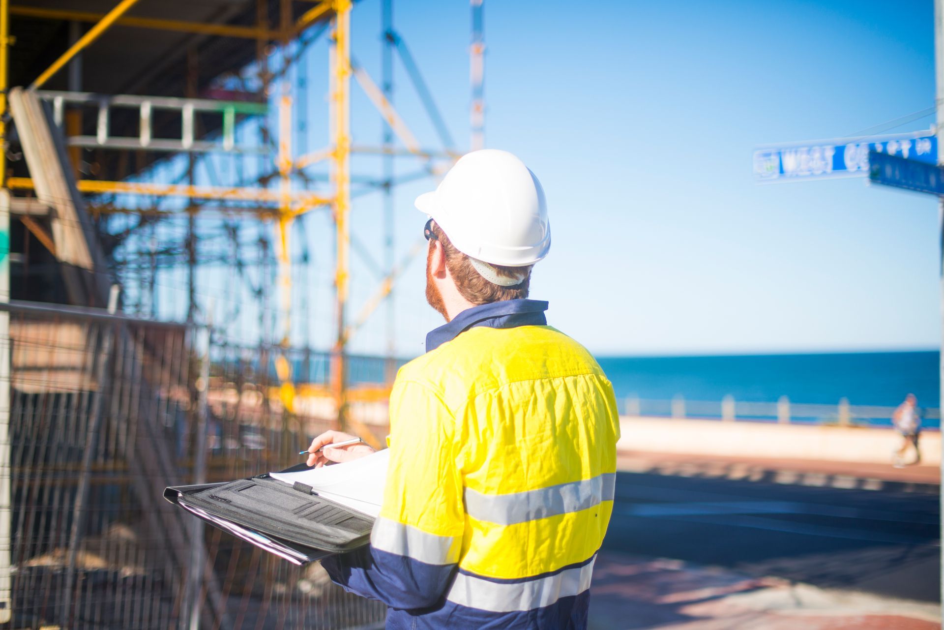 Construction worker in safety vest and hard hat inspects a site, writing on a clipboard; another worker in the background.