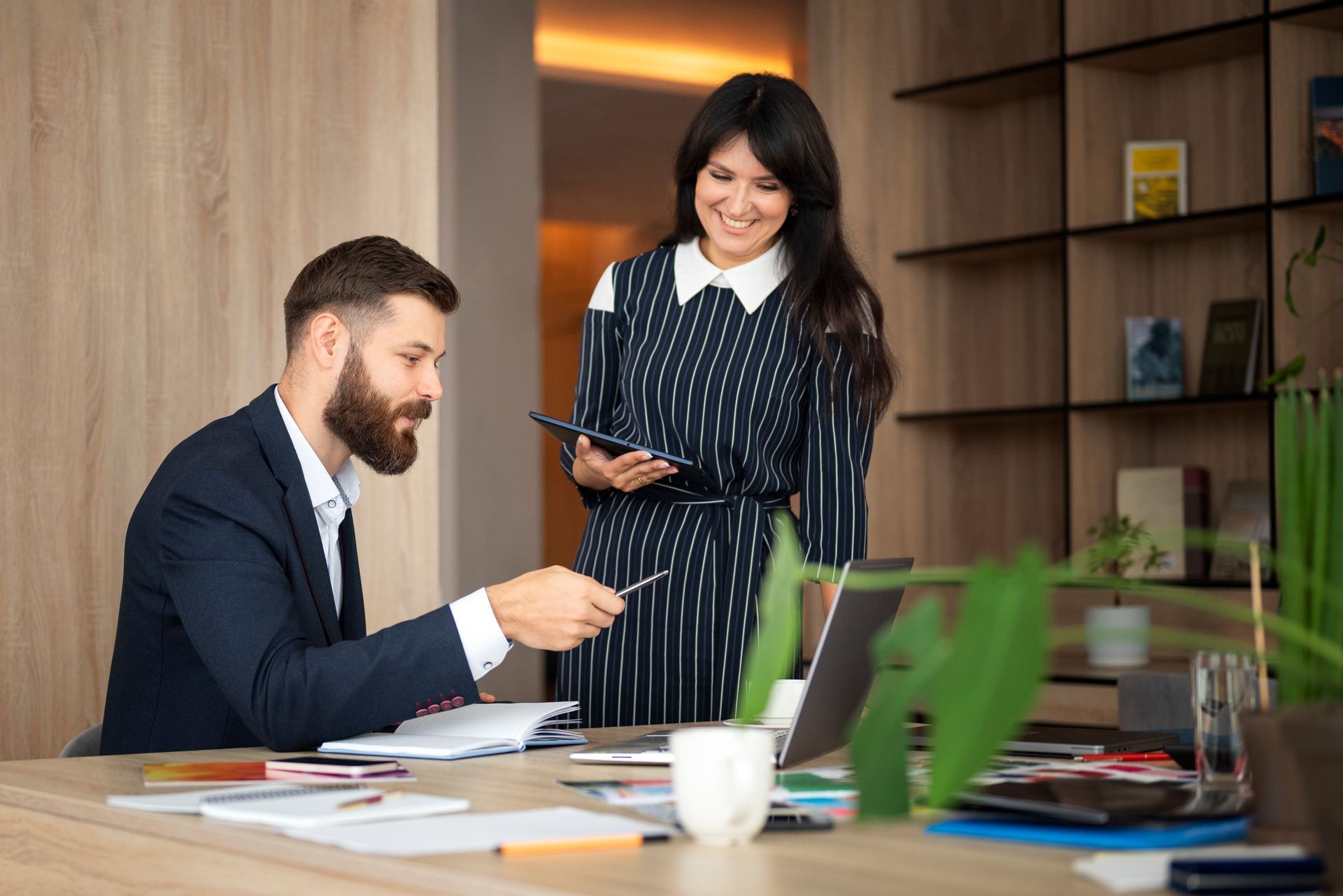 Man in suit points at documents, woman smiles, looking at tablet, office setting.