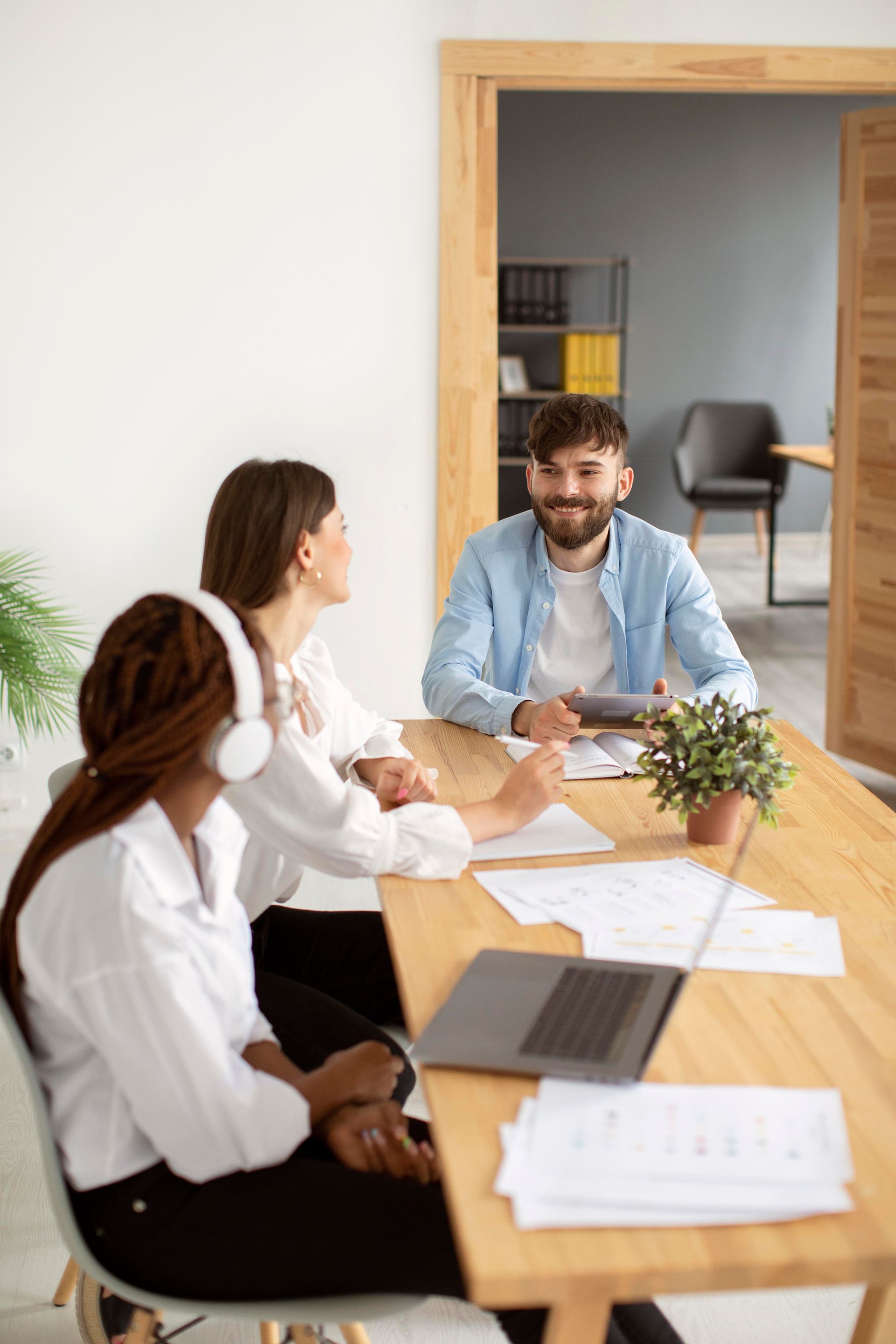 Three people at a table, discussing papers. Man smiles. Bright office setting, wood table.