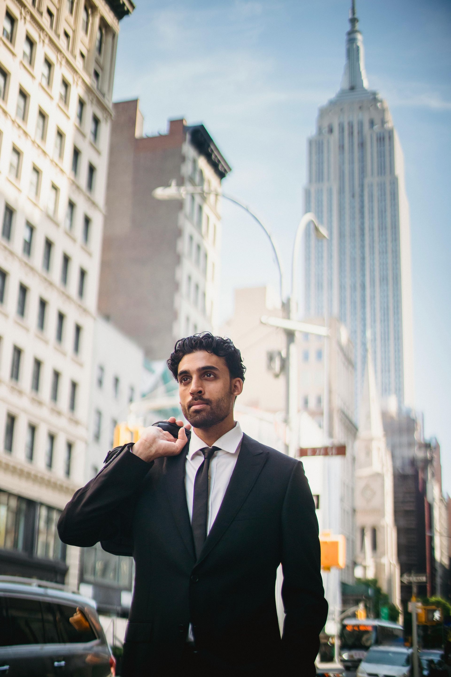 Man in suit, shoulder-bag, NYC street, Empire State Building in the background, looking upwards.