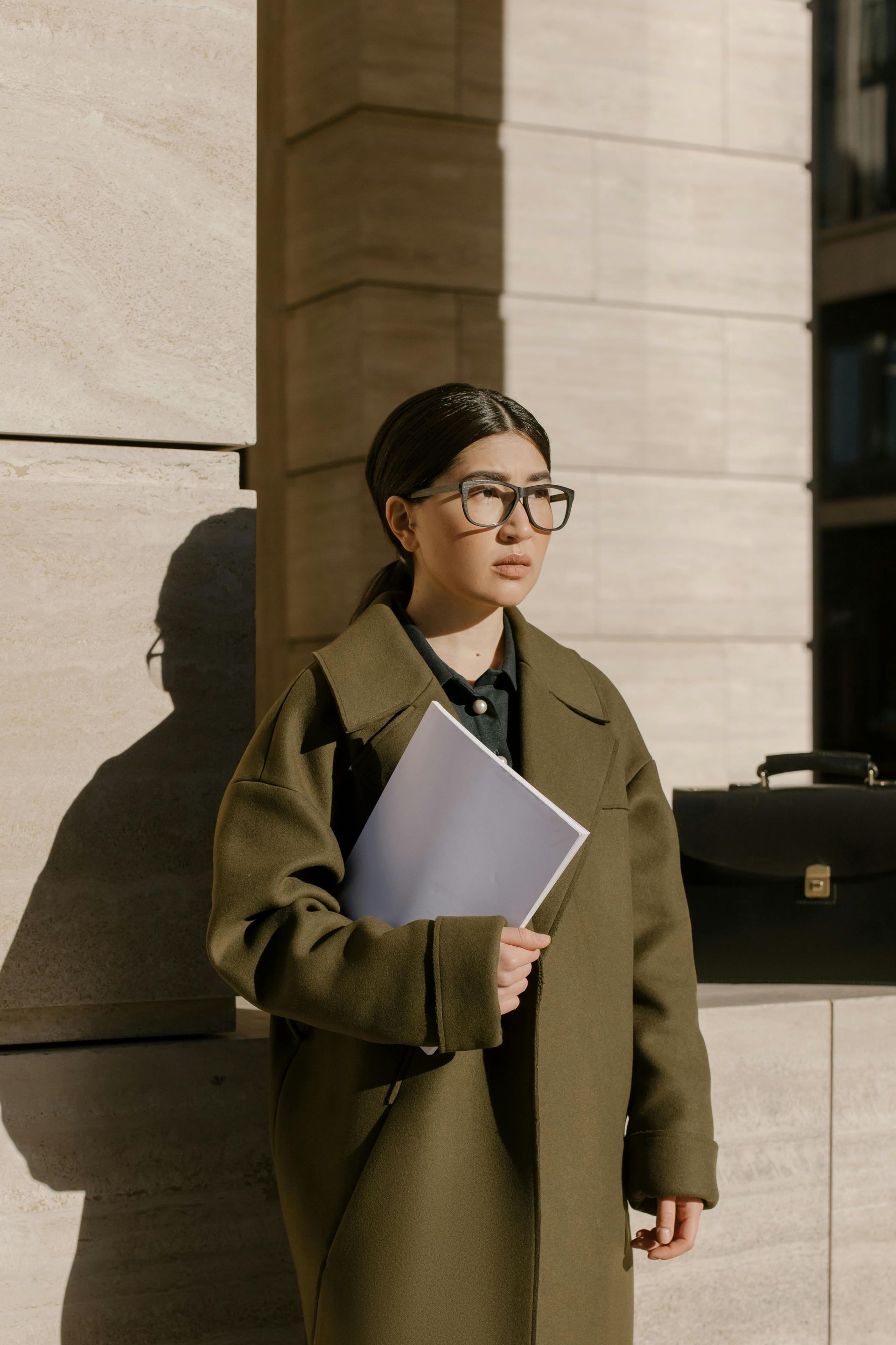 Woman in olive coat and glasses holds papers outside a building.