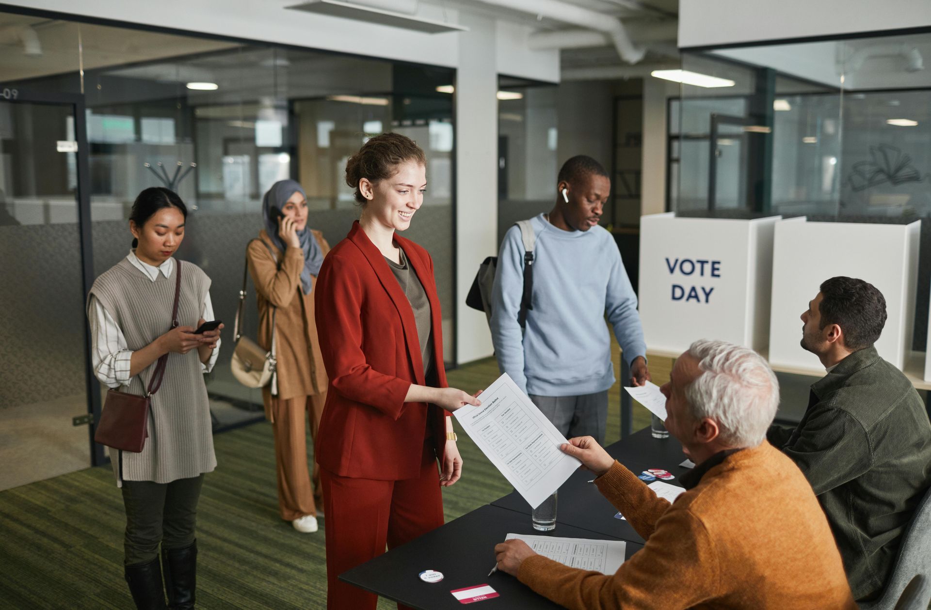 People voting at a polling place. A woman in a red suit hands a ballot to a poll worker.