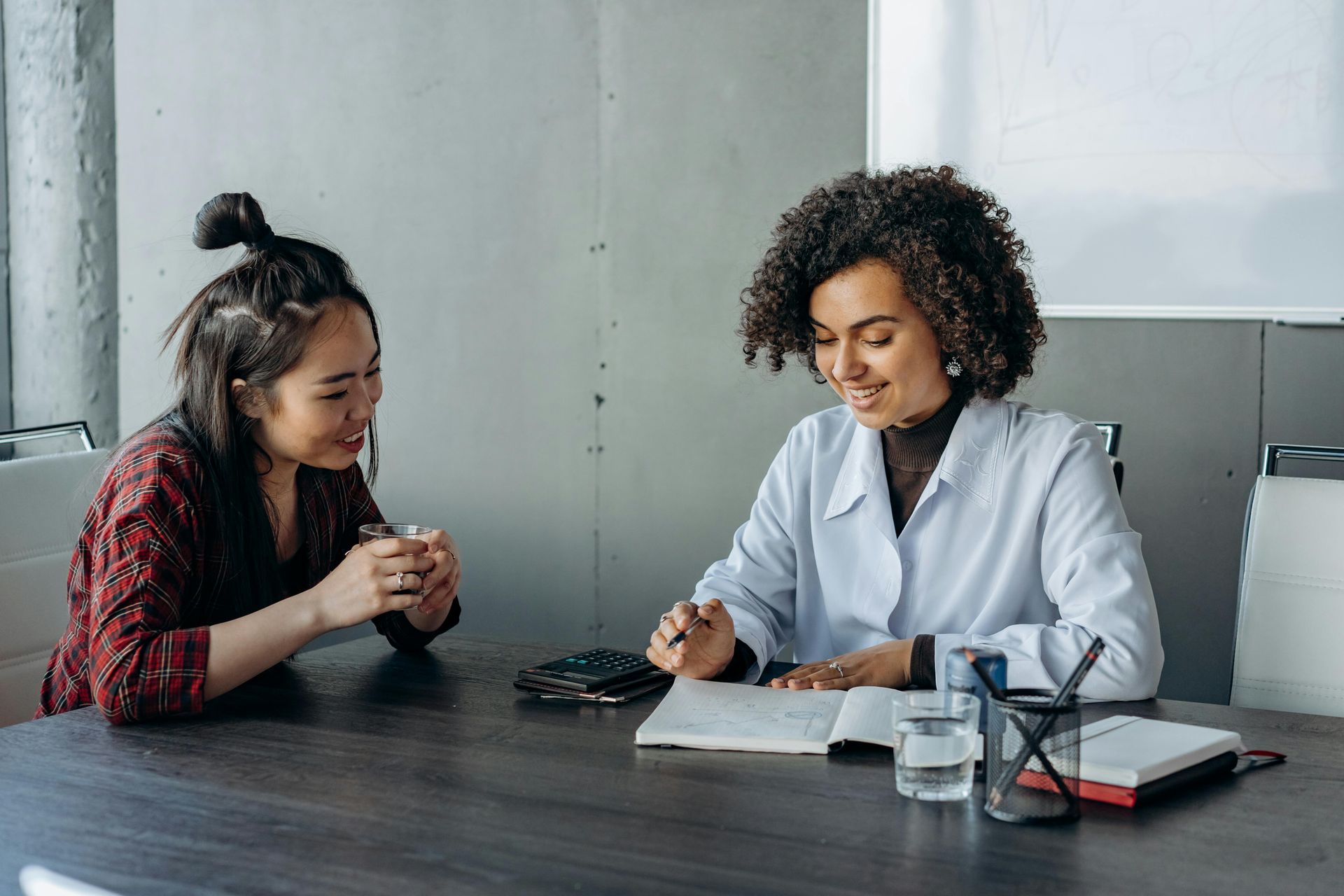 Two women seated at a desk, looking at a notebook. One is wearing a lab coat.